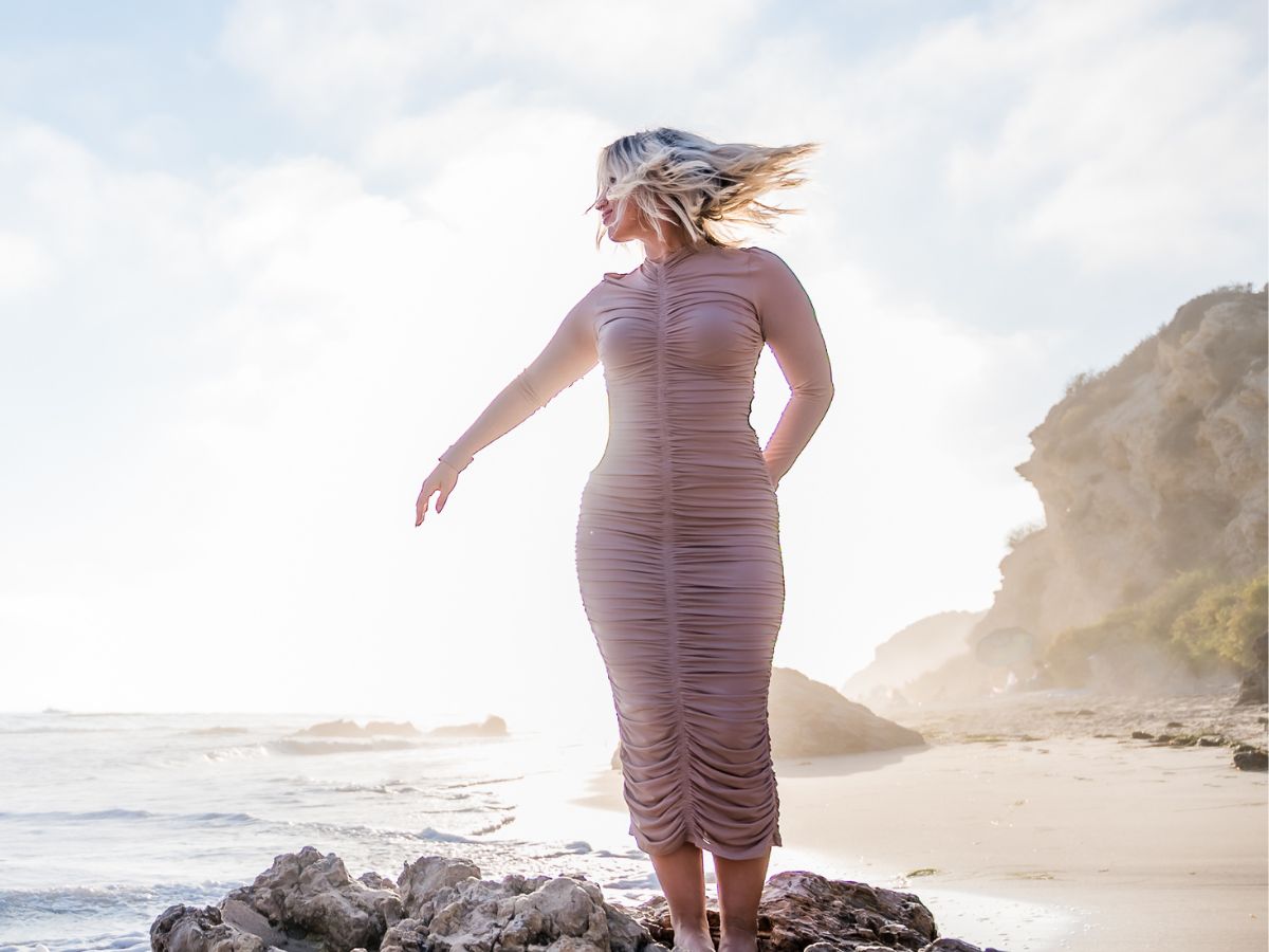 Cayla Craft standing confidently on rocks by the ocean, gazing into the distance with her hair blowing in the wind. She is wearing a fitted dress, embodying a sense of freedom, success, and abundance against the backdrop of a serene beach and clear sky, symbolizing growth and limitless possibilities.