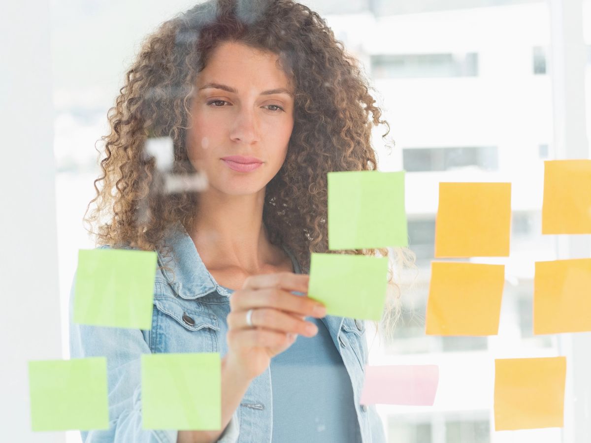 Young woman with curly hair, wearing a denim jacket, focusing and brainstorming with colorful sticky notes on a glass wall.