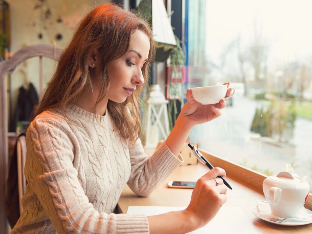 A woman sitting by the window in a cozy café, holding a cup of tea while thoughtfully reviewing notes, symbolizing reflection, planning, and the importance of taking time to focus on personal and financial growth.